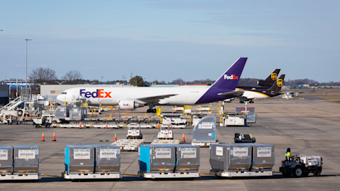 Aviones de carga de FedEx y UPS estacionados en la pista del aeropuerto con contenedores y vehículos de servicio al frente.