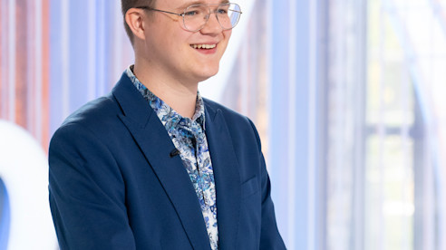 Person smiling and standing indoors wearing a blazer, with soft lighting and a modern background.