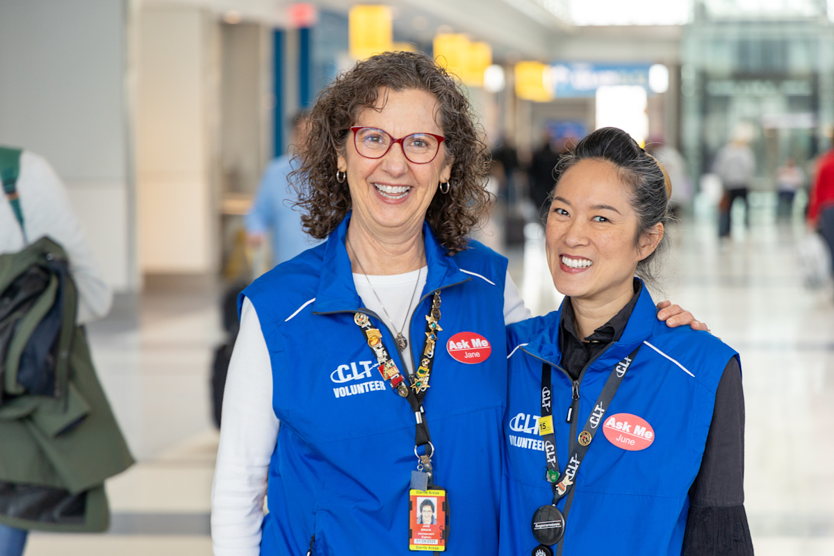 Two airport volunteers wearing blue “CLT Volunteer” vests smile while standing together in a terminal. Both wear red “Ask Me” name badges and ID lanyards. Travelers and airport signage appear in the background.