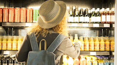 Una viajera con sombrero de paja y mochila elige una bebida de una vitrina refrigerada llena de botellas y latas.