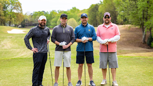 Four golfers standing on a golf course holding clubs, posing together on a grassy fairway.
