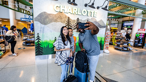 Travelers take a selfie in front of a Charlotte mural inside the airport, with luggage nearby.