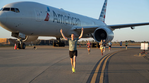 Runner raises arms while jogging past a parked airplane during an airport runway race event.
