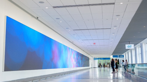 Viajeros pasan junto a una gran pared digital que muestra arte con tonos azules y púrpuras en un pasillo iluminado del aeropuerto.