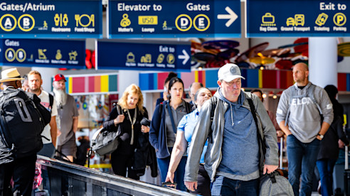 Passengers walk through a busy airport terminal under clear directional signage for gates, exits, and amenities.
