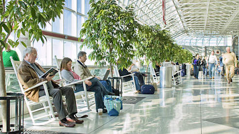 Viajeros se relajan en mecedoras a lo largo de un pasillo luminoso del aeropuerto con árboles y grandes ventanales.