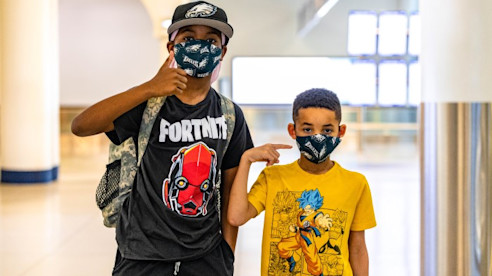 Dos niños con camisetas de personajes y mascarillas posan con el pulgar hacia arriba dentro de una terminal del aeropuerto.