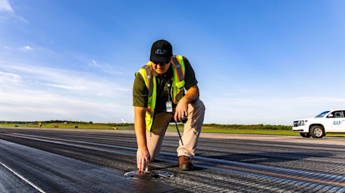 Un trabajador de operaciones del aeropuerto con chaleco de seguridad inspecciona una luz de pista en el campo de aviación bajo un cielo despejado.