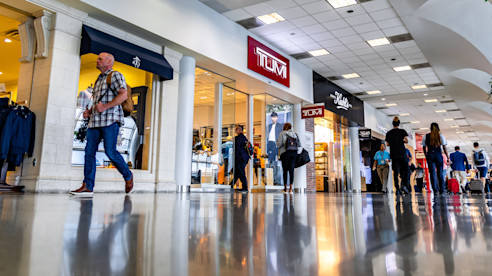 Viajeros caminan por un pasillo del aeropuerto rodeado de tiendas minoristas y vitrinas iluminadas.