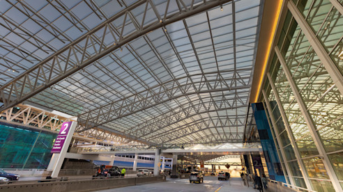Covered airport terminal drop-off with glass and steel canopy, cars, and passengers at curbside.
