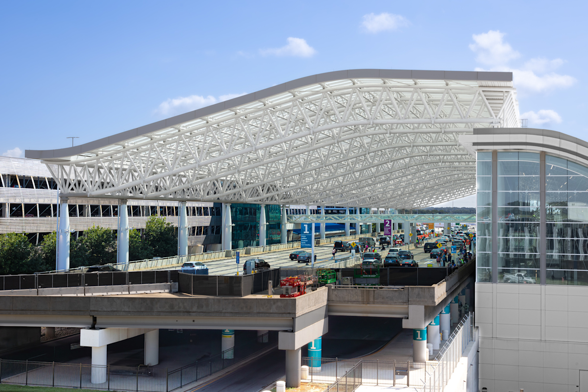 Vehicles pass through an airport terminal under a large white steel canopy with glass terminal structures.