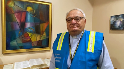 Un capellán del aeropuerto con chaleco de seguridad azul está de pie junto a una Biblia abierta y una pintura abstracta colorida.