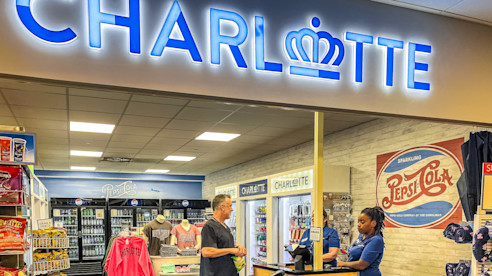 Airport convenience store with a bright "Charlotte" sign, stocked with snacks, drinks, and souvenirs, as a cashier assists a customer.