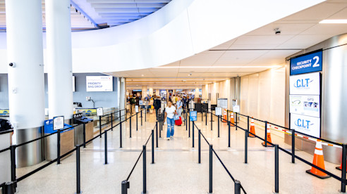 Airport security checkpoint with empty queue lines, digital screens, and a few passengers in the distance.