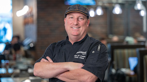 Chef en un restaurante posa con los brazos cruzados, sonriendo frente a un área de cocina.