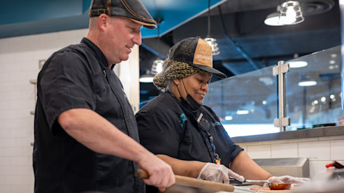 Dos empleados de cocina preparan alimentos juntos detrás de un mostrador en una cocina de restaurante.