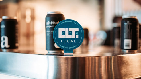Primer plano del tirador de cerveza CLT Local con latas negras de cerveza artesanal local al fondo en un bar del aeropuerto.
