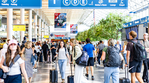 Busy airport concourse with travelers pulling luggage under blue overhead signs for gates and services.