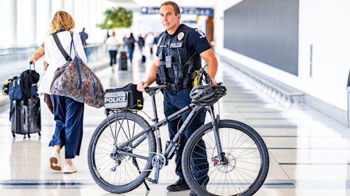 A police officer stands with a bicycle inside an airport concourse as travelers walk by with luggage.