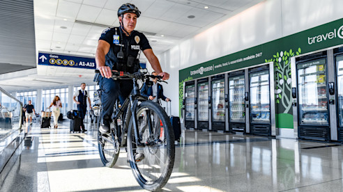 A police officer rides a bicycle through an airport concourse near vending machines and boarding gate signs.