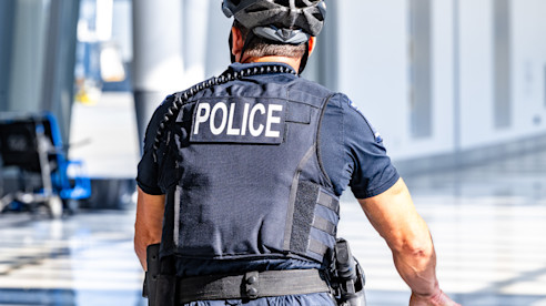 A police officer wearing a helmet and vest labeled “POLICE” walks with a bicycle inside the airport.