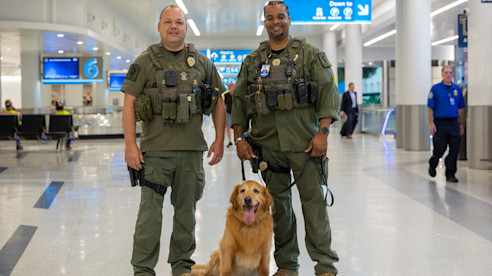 Dos oficiales uniformados posan en una terminal del aeropuerto con un perro K-9 sentado entre ellos.
