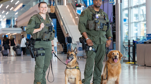 Dos oficiales uniformados están en una terminal del aeropuerto con sus perros K-9.