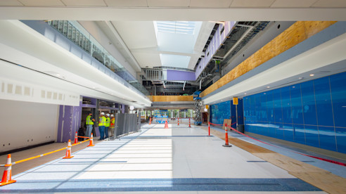 A modern airport concourse under construction, featuring spacious walkways, large skylights, and workers in high-visibility vests actively engaged in the project.