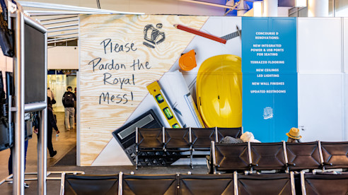 Construction wall inside airport with message “Please pardon the Royal mess!” and images of tools and hard hat.