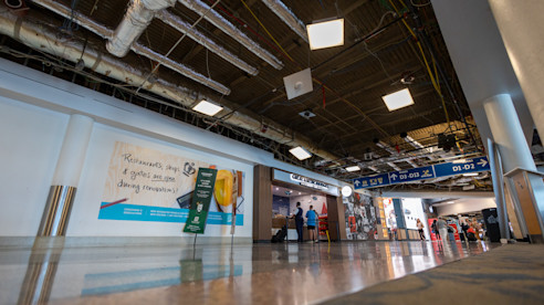 Airport hallway under construction with exposed ceiling pipes, signage for gates, and a renovation banner on the wall.