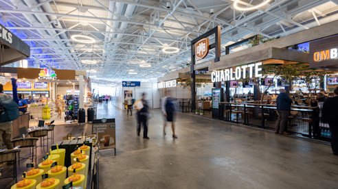 Airport concourse with shops, restaurants, and bright ceiling lights, including a bar with a Charlotte sign.