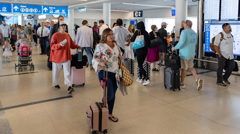Busy airport concourse with travelers pulling luggage near flight information displays and overhead signage.