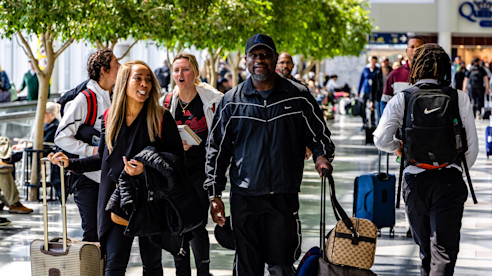 Travelers walk through an airport concourse with rolling luggage and indoor trees under bright lighting.