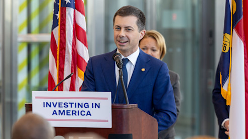 A public official speaks at a podium with an “Investing in America” sign, flanked by U.S. and state flags.