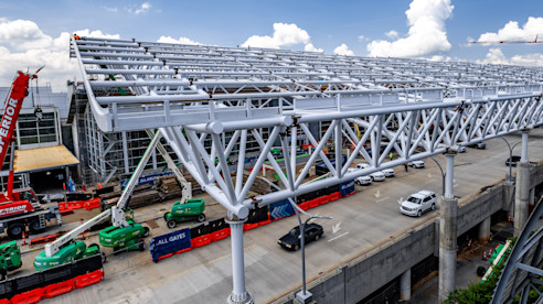 Steel truss canopy installation over an airport roadway with cranes, lifts, and construction vehicles below.