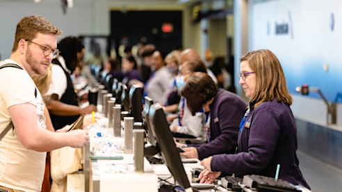Airport check-in counters with passengers assisted by airline staff at self-service kiosks.