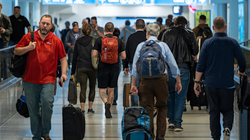 Travelers walking through a busy airport concourse with luggage and overhead directional signs.