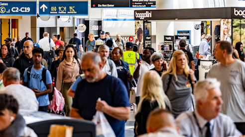 Busy airport concourse with travelers walking past shops and dining areas under overhead gate signage.
