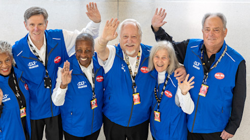 Group of airport volunteers wearing new blue uniforms smile and wave inside the terminal.