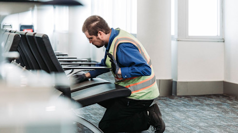 Un trabajador de mantenimiento del aeropuerto con chaleco de seguridad se arrodilla para inspeccionar o reparar los asientos en una sala de espera.