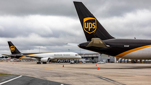 Dos aviones de carga de UPS están estacionados en la pista de un aeropuerto bajo un cielo nublado.