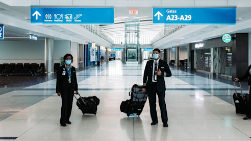 Miembros de la tripulación aérea con uniformes y mascarillas caminan por un pasillo vacío del aeropuerto con su equipaje.