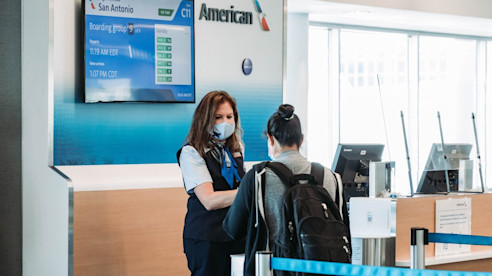 Agente de puerta de embarque ayuda a una viajera con mochila en el área de abordaje de American Airlines, con un vuelo a San Antonio mostrado en la pantalla.