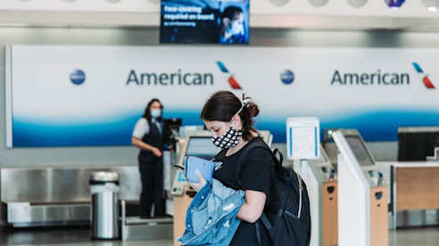 Una viajera con mascarilla y mochila revisa su teléfono cerca del mostrador de boletos de American Airlines.