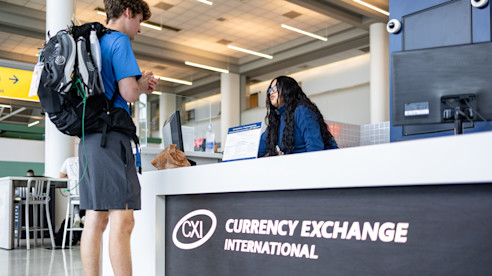 Viajero con mochila conversa con una agente en el mostrador de Currency Exchange International dentro del aeropuerto.