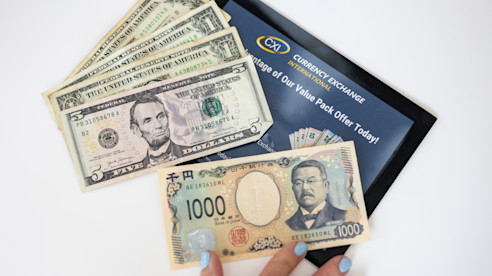 Mano sosteniendo un billete japonés de 1,000 yenes sobre varios billetes de dólares estadounidenses y un sobre de Currency Exchange International.