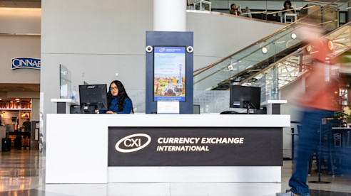 Mostrador de Currency Exchange International en el terminal del aeropuerto con una agente sentada detrás del escritorio y viajeros pasando.