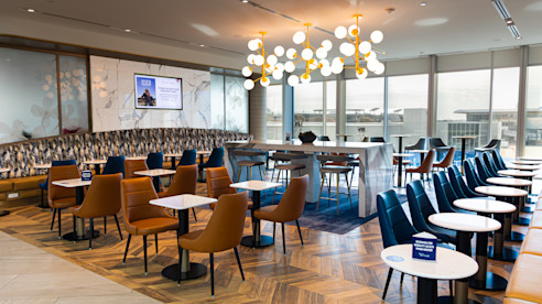 Modern airport lounge with stylish seating, marble tables, decorative lighting, and large windows overlooking the tarmac.