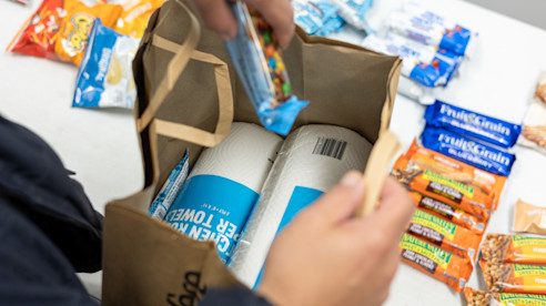 Manos colocan refrigerios y rollos de papel toalla en una bolsa de donaciones sobre una mesa interior.