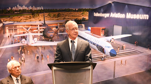 Man speaking at a podium inside an aviation museum with aircraft exhibits and a mural in the background.